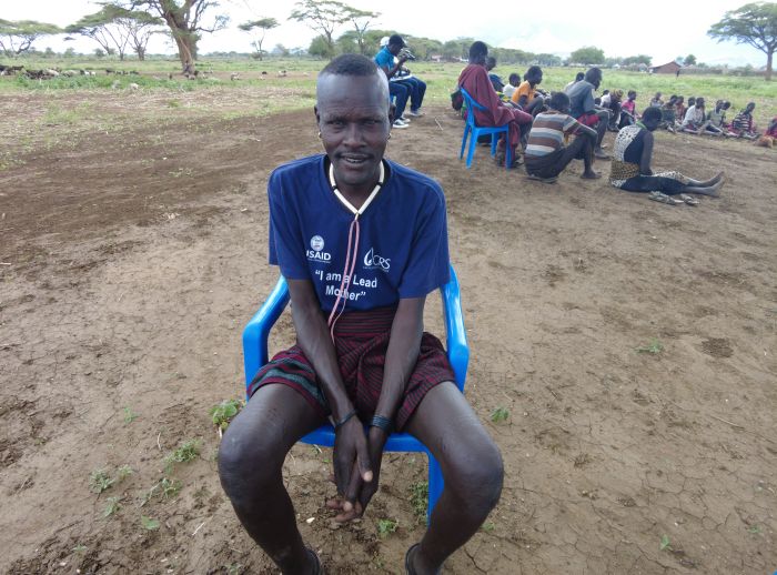 Nangiro David sitting on a blue plastic chair next to an ongoing outdoor farming class. 