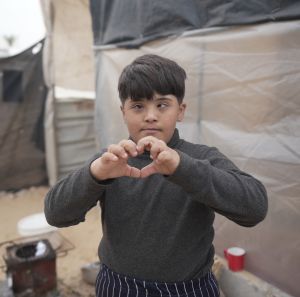 Image of a kid in a camp in Gaza forming a heart with his hand.