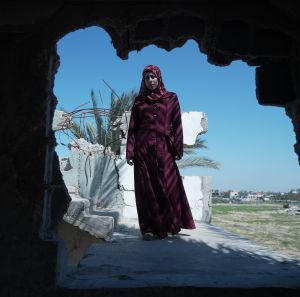 A woman in a red dress stands in a bombed-out building, symbolizing maternal hardship in Gaza, supported by the Power to Choose Programme.