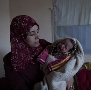 Suhad holds her child in a room with red chairs and a curtained window